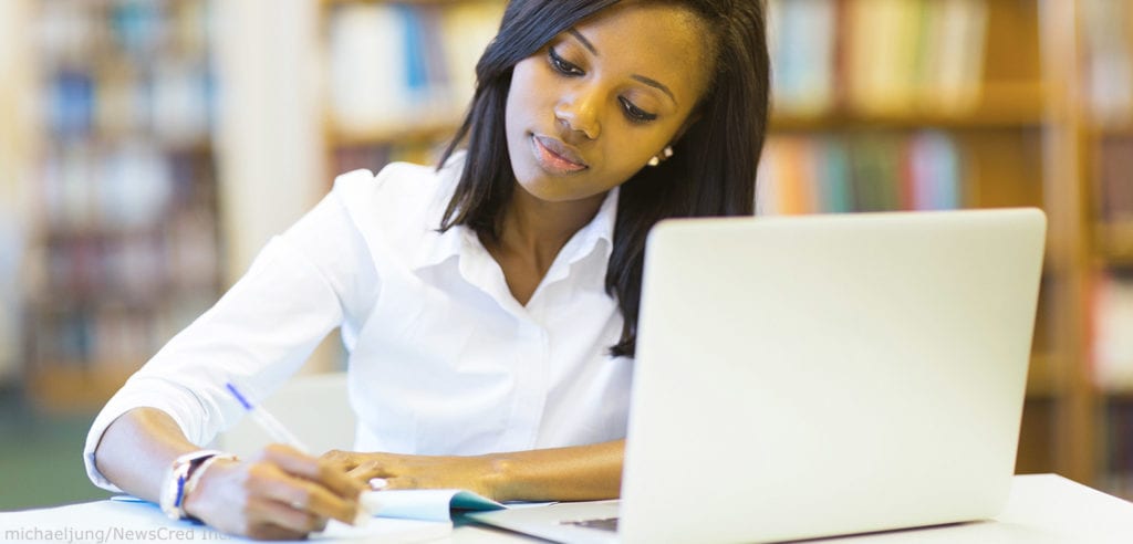 Female African American college student writing in a notebook with a laptop